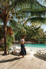 Wall Mural - Young woman in straw hat walk near the swimming pool under palm tree. Tropical summer vacation concept