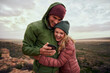 © StratfordProductions - Laughing young couple embracing during camp on mountains looking at smartphone in winter clothing