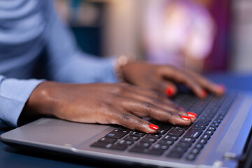 Close up of dark skinned business woman hand typing email n laptop computer from home office late at night. Employee using modern technology network wireless doing overtime.