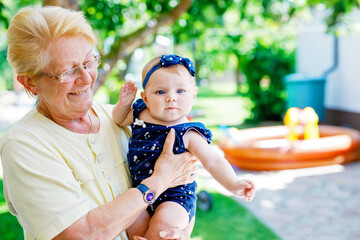 Wall Mural - Cute little baby girl with grandmother on summer day in garden. Happy senior woman holding smiling child on arm