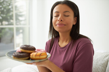 Close up shot of beautiful young dark skinned woman having pleased facial expression keeping her eyes closed smelling sweet scent of delicious colorful doughnuts on plate, smiling in enjoyment