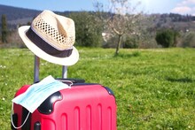 Light Blue Surgical Mask On Luggage And A Hat, In A Beautiful European Green Background. Face Mask Is Compulsory For Travelers, New Normal.