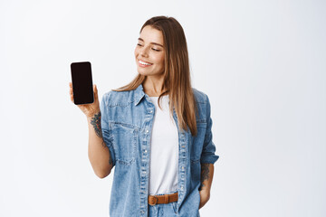 Stylish girl showing empty mobile phone screen and looking pleased, recommending application for smartphone, demonstrating shopping site, white background