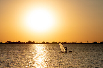 Woman windsurfer silhouette at lake sunset. Beautiful beach landscape. Summer water sports activities, recreation and travel concept