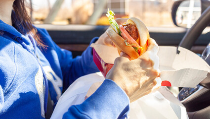 A woman is eating chicken sandwich which she got from a roadside drive through fast food chain. She has packs of food on her lap as she eats in the driver seat of her car in the parking lot.