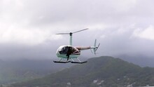 Slow Motion Close-up Of A Helicopter Hovering Over The Sea Surface With A Photographer Hanging Outside The Side Door Under Cloudy Skies And With Steep Mountains In The Background - Oahu, Hawaii