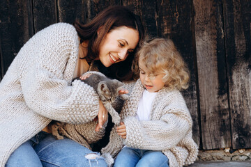 mother and her daughter play with a little cat by the wooden wall.