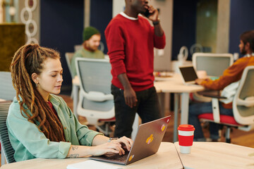 Poster - Young Caucasian businesswoman with dreadlocks sitting by desk in front of laptop