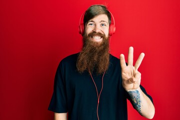 Canvas Print - Redhead man with long beard listening to music using headphones showing and pointing up with fingers number three while smiling confident and happy.