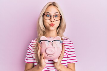 Sticker - Beautiful caucasian blonde girl holding piggy bank with glasses puffing cheeks with funny face. mouth inflated with air, catching air.