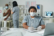 © pressmaster - Young mixed-race clinician in protective workwear filling in medical document by desk