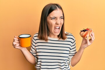 Poster - Young beautiful woman drinking coffee and eating pastry angry and mad screaming frustrated and furious, shouting with anger. rage and aggressive concept.