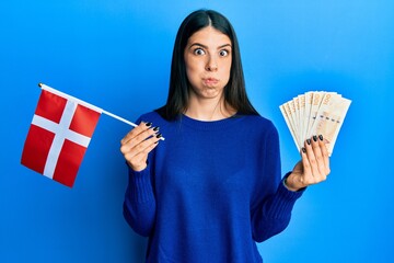 Young hispanic woman holding denmark flag and krone banknotes puffing cheeks with funny face. mouth inflated with air, catching air.