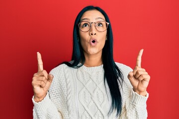 Sticker - Beautiful hispanic woman wearing casual sweater and glasses amazed and surprised looking up and pointing with fingers and raised arms.