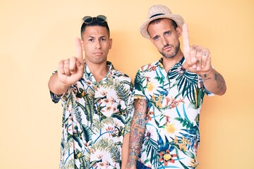 Wall Mural - Young gay couple of two men wearing summer hat and hawaiian shirt pointing with finger up and angry expression, showing no gesture