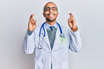 Wall Mural - Hispanic adult man wearing doctor uniform and stethoscope gesturing finger crossed smiling with hope and eyes closed. luck and superstitious concept.