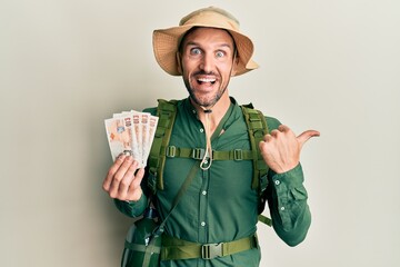 Wall Mural - Handsome man with beard wearing explorer hat holding 10 pounds pointing thumb up to the side smiling happy with open mouth