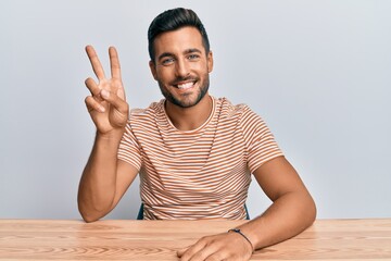 Handsome hispanic man wearing casual clothes sitting on the table showing and pointing up with fingers number two while smiling confident and happy.