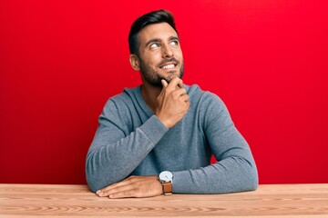 Wall Mural - Handsome hispanic man wearing casual style sitting on the table with hand on chin thinking about question, pensive expression. smiling with thoughtful face. doubt concept.