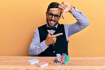 Sticker - Handsome hispanic croupier man sitting on the table with poker chips and cards smiling making frame with hands and fingers with happy face. creativity and photography concept.