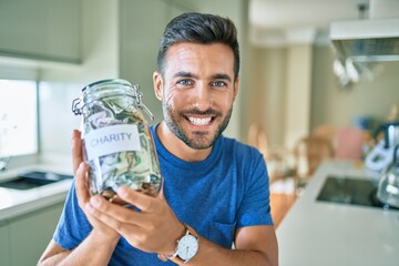 Young handsome man smiling happy holding charity jar with money at home