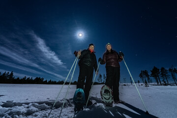 Beautiful middle aged men and women in snowshoes stand in night rare snowy winter forest under full moon light. Night walk, Lapland, Umea, Sweden