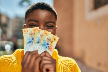 Poster - Young african american woman covering face with swiss franc banknotes at the city.