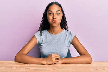 Poster - Young african american girl wearing casual clothes sitting on the table puffing cheeks with funny face. mouth inflated with air, crazy expression.