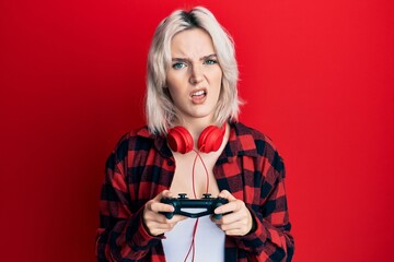 Sticker - Young blonde girl playing video game holding controller in shock face, looking skeptical and sarcastic, surprised with open mouth