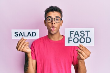 Sticker - Young handsome african american man holding salad and fast food message paper making fish face with mouth and squinting eyes, crazy and comical.