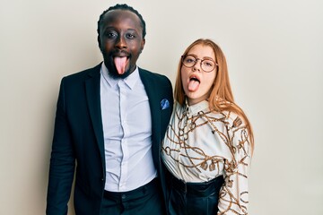 Wall Mural - Young interracial couple wearing business and elegant clothes sticking tongue out happy with funny expression. emotion concept.