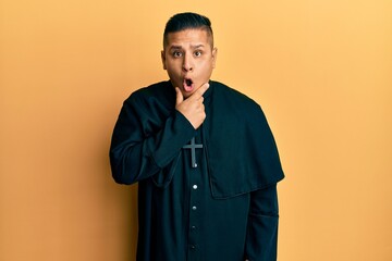 Canvas Print - Young latin priest man standing over yellow background looking fascinated with disbelief, surprise and amazed expression with hands on chin