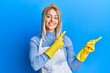 © Krakenimages.com - Beautiful blonde woman wearing cleaner apron and gloves smiling and looking at the camera pointing with two hands and fingers to the side.