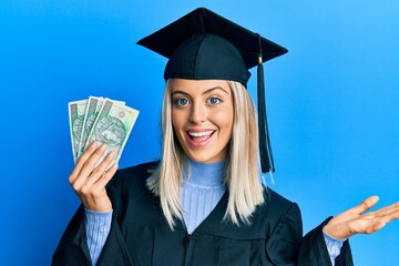 Beautiful blonde woman wearing graduation cap and ceremony robe holding polish zloty celebrating achievement with happy smile and winner expression with raised hand