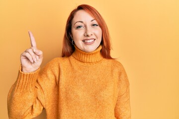 Poster - Beautiful redhead woman wearing casual winter sweater over yellow background with a big smile on face, pointing with hand finger to the side looking at the camera.