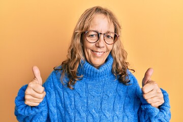 Middle age blonde woman wearing glasses and casual winter sweater success sign doing positive gesture with hand, thumbs up smiling and happy. cheerful expression and winner gesture.