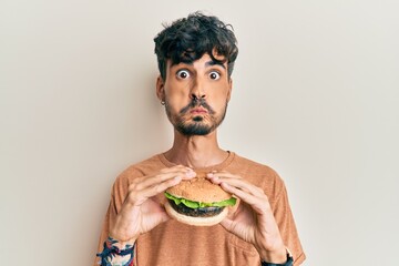 Poster - Young hispanic man eating a tasty classic burger puffing cheeks with funny face. mouth inflated with air, catching air.