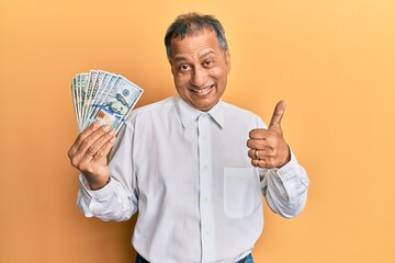 Sticker - Middle age indian man holding dollars smiling happy and positive, thumb up doing excellent and approval sign