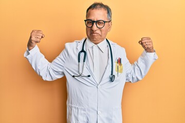 Canvas Print - Middle age indian man wearing doctor coat and stethoscope showing arms muscles smiling proud. fitness concept.