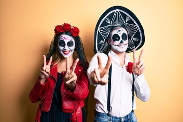 Wall Mural - Couple wearing day of the dead costume over yellow smiling looking to the camera showing fingers doing victory sign. number two.