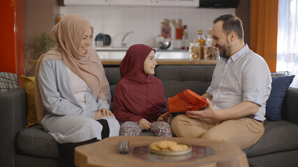 The affectionate little girl in hijab gives the gift box she bought with her mother to her father on father's day. Young father hugs his little daughter and kisses. 