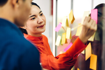 Casual creative happy two smile asian business people planning strategy analysis and brainstorm putting post it stickers note to share idea for startup project on glass board at modern office