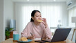© ChayTee - Young attractive beautiful asian female hungry eat doughnut take away snack food with full mouth look at computer notebook at home in busy work from home multitask unhealthy meal lifestyle concept.