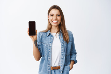 Portrait of attractive young woman with white smile, showing empty smartphone screen, demonstrate an app or online shopping store, studio background