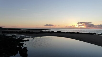 Wall Mural - 4k dynamic aerial video of sunset on a sand beach with shallow ponds of water, poles, rocks and small pier