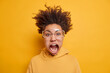 Emotional outbreak concept. Crazy shocked dark skinned woman screams excited looks in terror at camera being super angry and outraged wears big transparent glasses and sweatshirt isolated on yellow