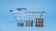 Coin stacks in front of empty shopping basket on blue background. Shopping expenses and retail growth concept