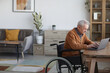 Side view portrait of senior man in wheelchair using laptop while working at desk in home interior, copy space