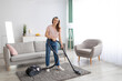 Happy young housemaid tidying her flat, vacuuming rug, indoors. Professional cleaning service concept