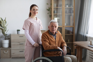 portrait of young female nurse looking at camera while helping senior man in wheelchair, copy space